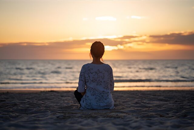 夕陽を見ながら、海辺の砂浜に座っている女性の後ろ姿