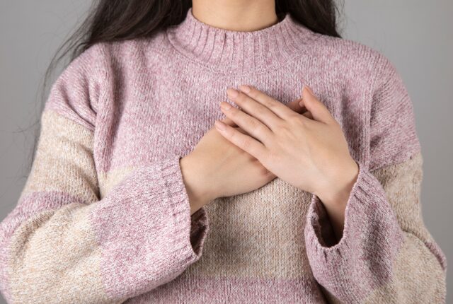 ピンク色のセーターを着た黒髪ロングヘアの女性、両手を心臓付近の胸に当てている